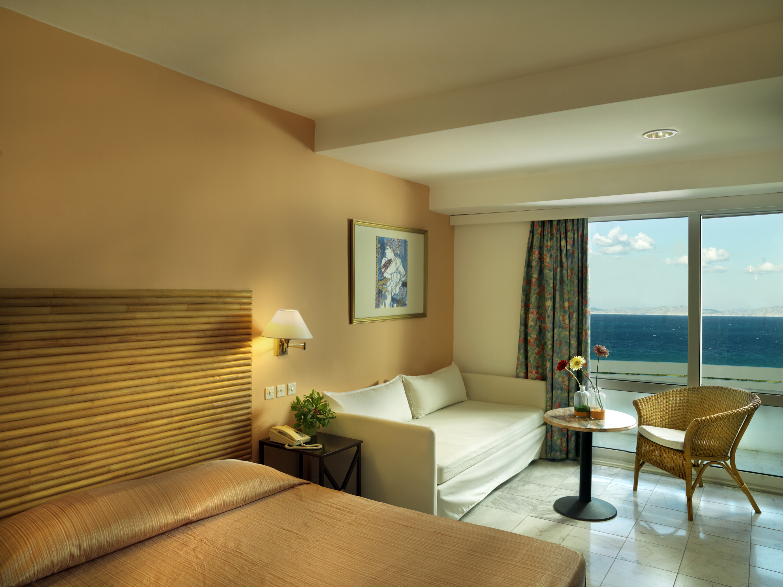Γοητευτική εικόνα ενός δωματίου στο ξενοδοχείο Dionysos, με το κρεβάτι, το καθιστικό και το μπαλκόνι με θέα στη θάλασσα.